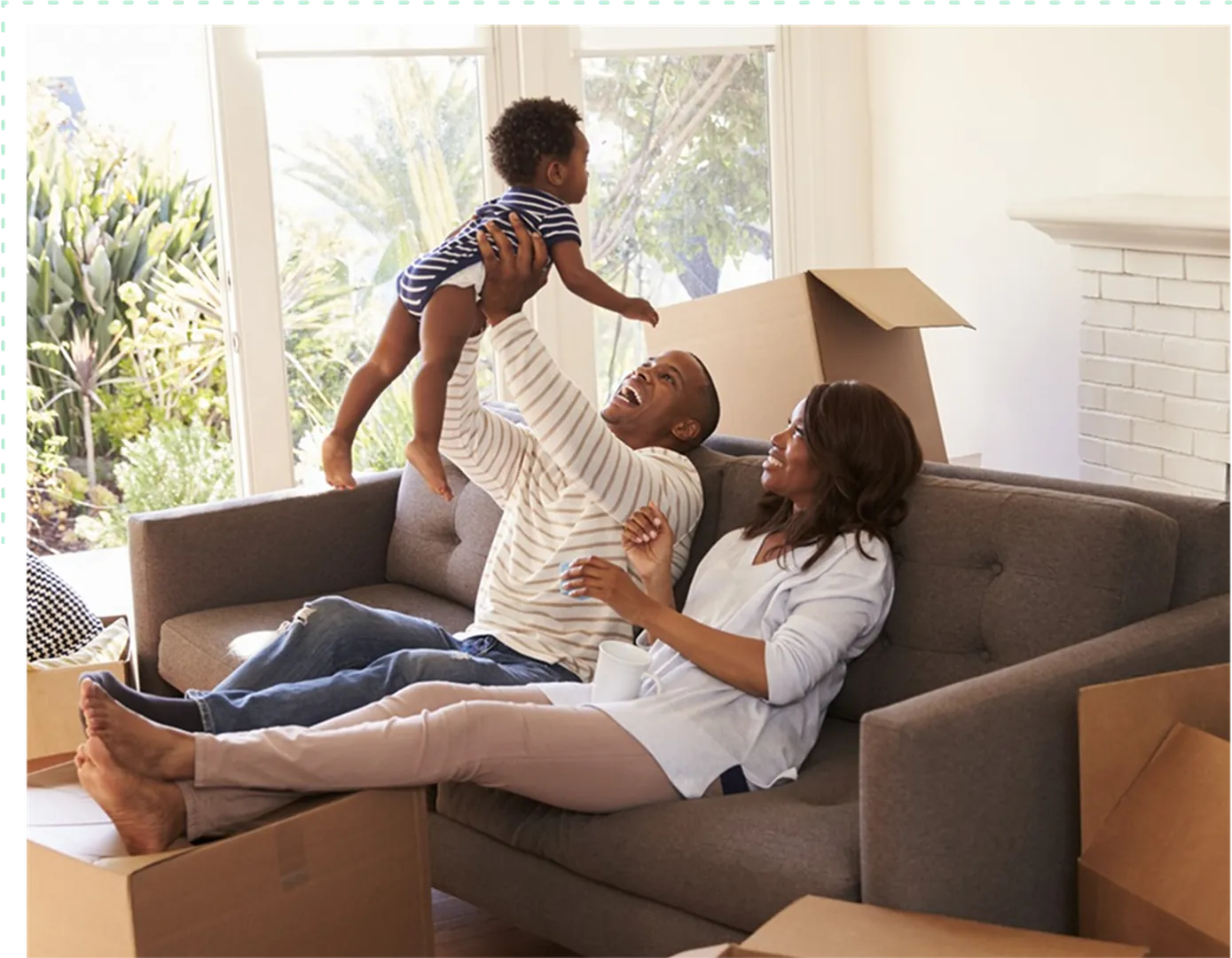 A joyful family plays together on a couch, holding their child, surrounded by moving boxes.