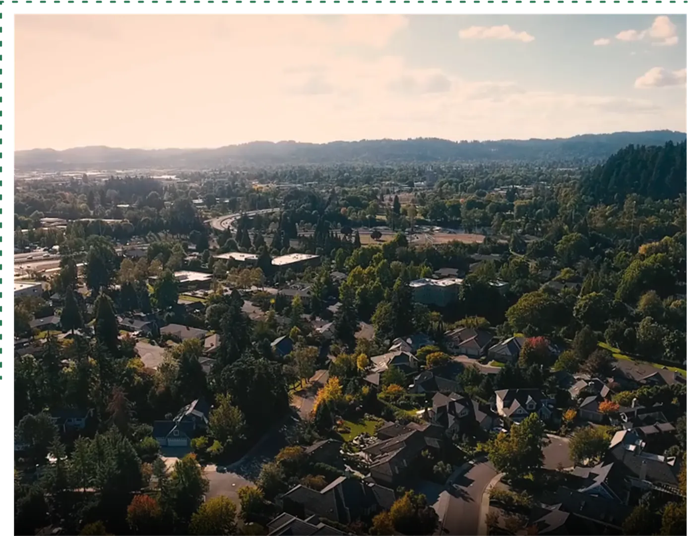 Aerial view of a lush suburban neighborhood surrounded by trees and hills, bathed in warm sunlight.