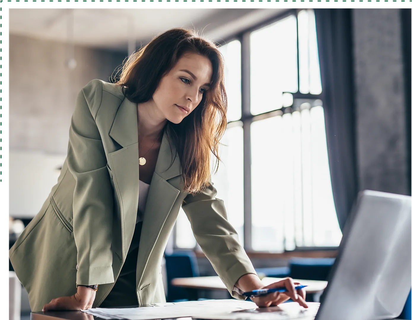 A woman in a green blazer leans over a table, writing with a pen, in a workspace with large windows.
