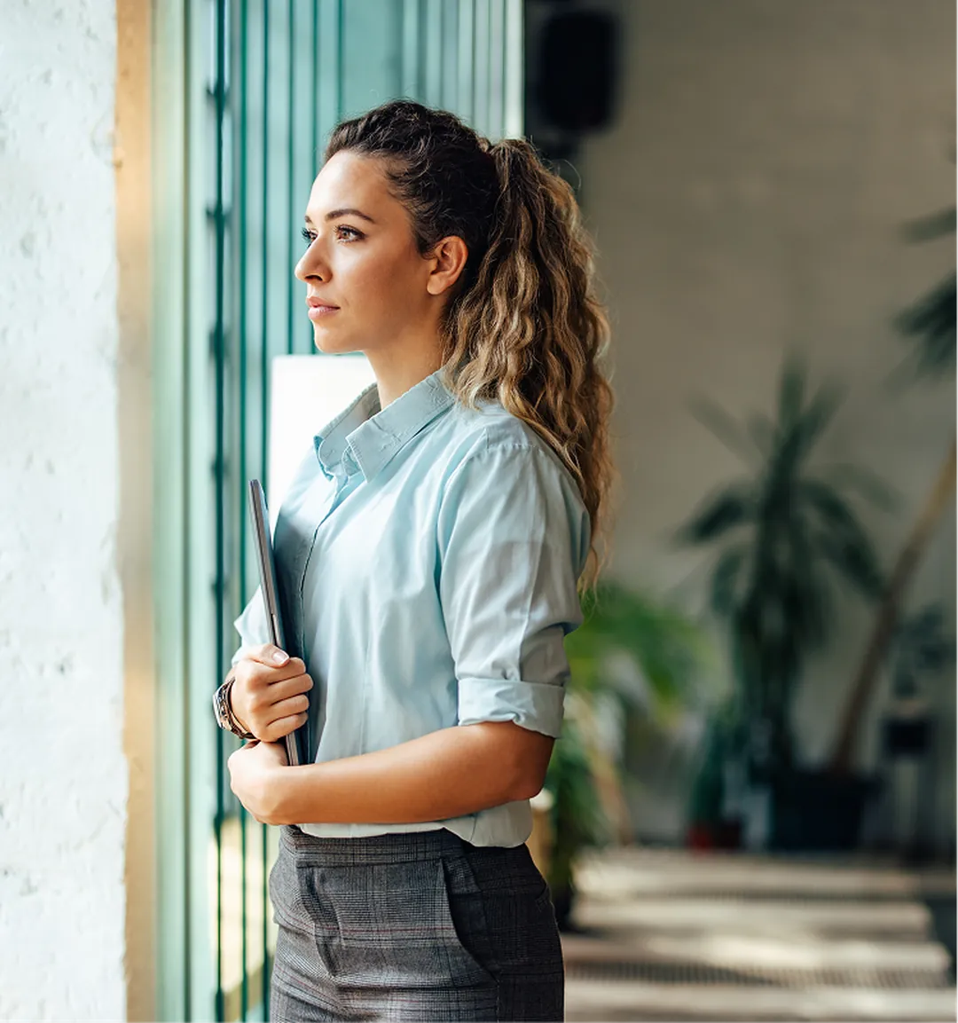 Woman with long curly hair holding files, standing by a window and looking into the distance.