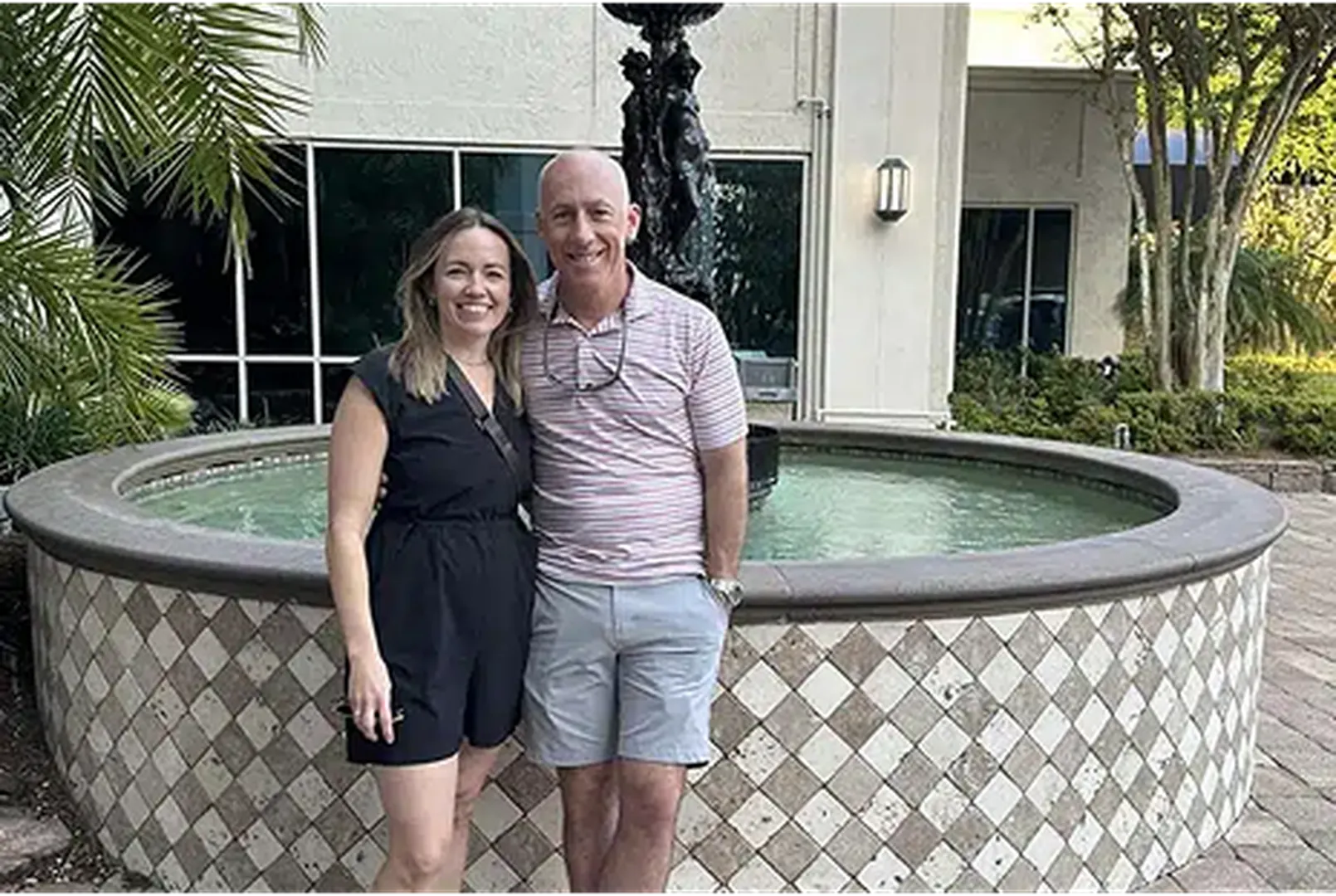 A couple stands next to a decorative fountain surrounded by lush greenery and palm leaves.