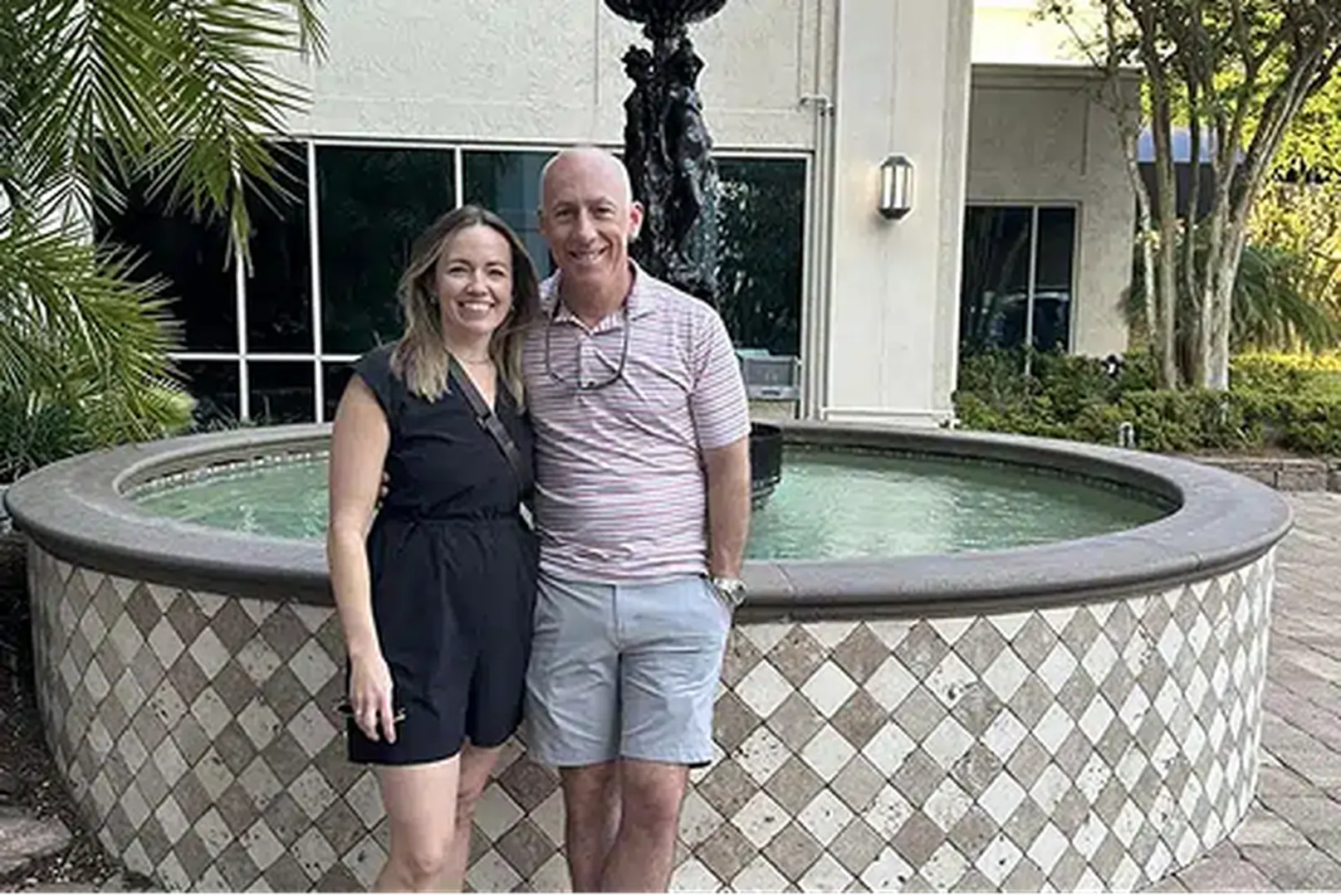 A couple stands next to a decorative fountain surrounded by lush greenery and palm leaves.