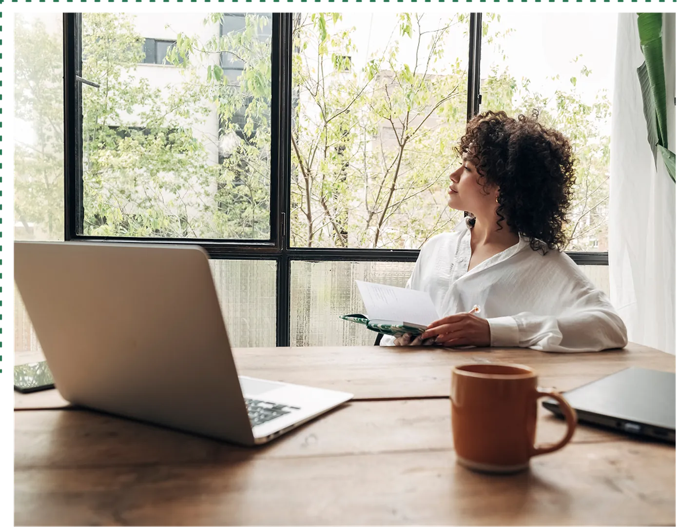 A woman with curly hair in a white shirt sitting at an office desk with a large window behind.
