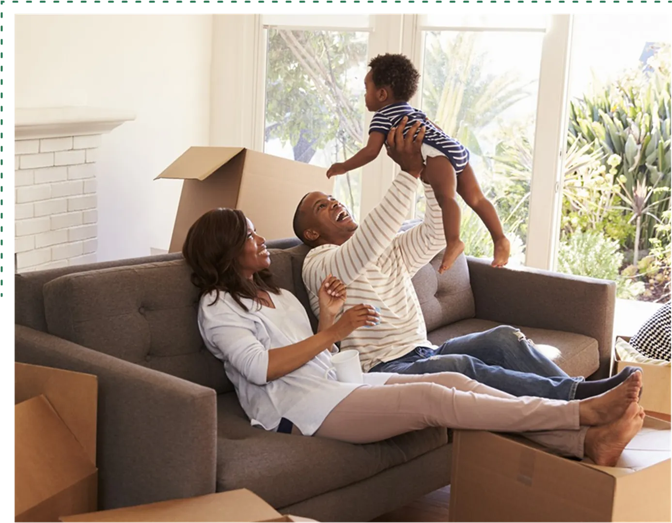 A couple playing on a couch, holding their child, having a large window behind and boxes around.